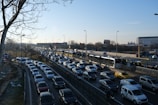 Heavy traffic is seen on a multi-lane road during daylight. A long line of cars is closely packed, suggesting congestion. On the right side, buses are using dedicated lanes. Sparse trees are visible on the left, with leafless branches indicating winter or early spring. Several billboard advertisements and a large building can be seen in the background.