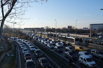 Heavy traffic is seen on a multi-lane road during daylight. A long line of cars is closely packed, suggesting congestion. On the right side, buses are using dedicated lanes. Sparse trees are visible on the left, with leafless branches indicating winter or early spring. Several billboard advertisements and a large building can be seen in the background.