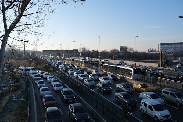 Heavy traffic is seen on a multi-lane road during daylight. A long line of cars is closely packed, suggesting congestion. On the right side, buses are using dedicated lanes. Sparse trees are visible on the left, with leafless branches indicating winter or early spring. Several billboard advertisements and a large building can be seen in the background.
