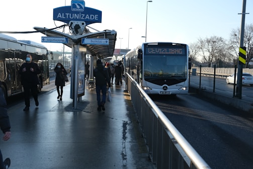 A bus stop at Altunizade with several people walking and waiting. Two buses are parked, and a sign indicates directions to Sogutlucesme and Beylikduzu. The sky is clear with bare trees in the background, and the ground appears wet.