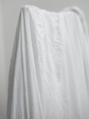 A white fabric draped over an object, creating soft folds and shadows. The texture of the material is visible, showing creases and slight wrinkles. There is a neutral, minimalist background.