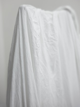 A white fabric draped over an object, creating soft folds and shadows. The texture of the material is visible, showing creases and slight wrinkles. There is a neutral, minimalist background.