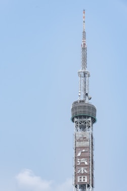 A tall, slender telecommunications tower featuring a lattice structure and a circular platform near the top. The tower includes signage with visible Chinese characters. The background is a clear blue sky.