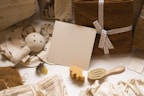 Various baby items are arranged neatly, including a soft plush bunny toy, a small wooden elephant, a baby hairbrush, and a pacifier. There are also folded blankets and towels, some tied with a ribbon, and fabric with cherry patterns. A blank beige card is placed centrally among these items.