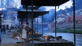 A man is seen tending to a cremation pyre by a riverbank, surrounded by structures with corrugated metal roofs. Smoke rises from the pyre, blending with the twilight sky. In the background, there are people walking along the riverbank and buildings with traditional architectural features.