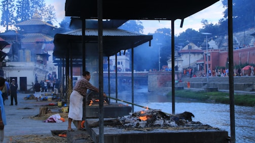 A man is seen tending to a cremation pyre by a riverbank, surrounded by structures with corrugated metal roofs. Smoke rises from the pyre, blending with the twilight sky. In the background, there are people walking along the riverbank and buildings with traditional architectural features.
