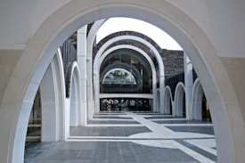 A series of white stone arches creating an architectural corridor, leading towards a modern building facade with glass elements. The arches form a repeating geometric pattern, enhancing the perspective and depth within the space.