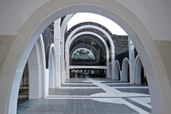 A series of white stone arches creating an architectural corridor, leading towards a modern building facade with glass elements. The arches form a repeating geometric pattern, enhancing the perspective and depth within the space.