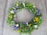 A circular floral wreath composed of a variety of fresh flowers and green leaves is placed on a concrete surface. The flowers include daisies, yellow button flowers, purple blooms, and small white blossoms, creating a colorful and vibrant arrangement.