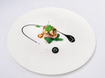 A minimalist and artistic food presentation on a large white plate featuring a small portion of meat garnished with herbs and mushrooms. Dark sauce swirls artfully around the dish, adding an elegant touch.
