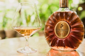 A large bottle of Rémy Martin Premier Cru cognac is positioned upright on a reflective surface. Next to it, there is an elegant brandy snifter containing a small amount of the amber liquid. The background is softly blurred with hints of green foliage, creating a serene and luxurious setting.