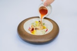 A bowl of artistically arranged food sits in a rustic, brown-edged ceramic bowl. The dish contains an assortment of colorful ingredients including greens, sliced radish, and a pale, creamy component. A hand is seen pouring a vibrant orange sauce from a small white pitcher onto the food.
