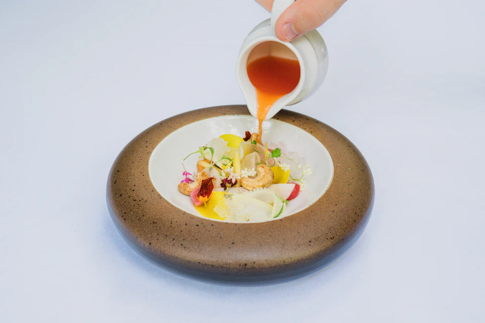 Hand pours sauce onto a plate with various food items, including shrimp and vegetables