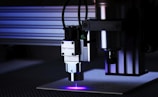 A close-up view of a laser engraver in action. The machine has a metallic build with cables attached and is engraved with the label 'Opt Lasers'. The laser is operating, emitting a blue laser beam onto the surface below.