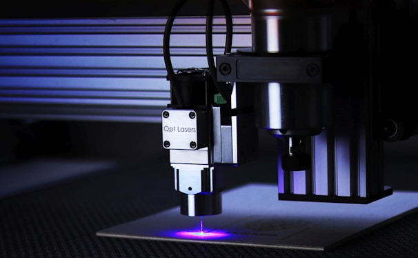 A close-up view of a laser engraver in action. The machine has a metallic build with cables attached and is engraved with the label 'Opt Lasers'. The laser is operating, emitting a blue laser beam onto the surface below.