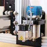 A close-up view of a CNC machine or router setup equipped with a laser module. The machine features a metallic frame with wheels for linear motion, while the blue component likely houses a motor or other rotary tool. The laser module is clearly marked with a warning symbol, indicating its functionality.