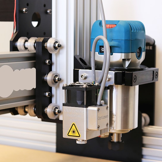 A close-up view of a CNC machine or router setup equipped with a laser module. The machine features a metallic frame with wheels for linear motion, while the blue component likely houses a motor or other rotary tool. The laser module is clearly marked with a warning symbol, indicating its functionality.