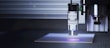 A precision laser cutting machine appears in a workspace, with a laser head marked 'Opt Lasers' actively cutting or engraving material. The machine is mounted on a linear rail system, and the surrounding area is dark, highlighting the blue light emitted by the laser.