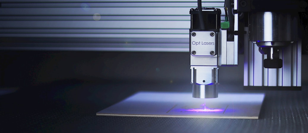 A precision laser cutting machine appears in a workspace, with a laser head marked 'Opt Lasers' actively cutting or engraving material. The machine is mounted on a linear rail system, and the surrounding area is dark, highlighting the blue light emitted by the laser.
