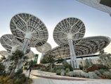 Innovative, large solar panel structures designed like futuristic trees, situated in a landscaped area with various plants and a pathway. The scene includes people walking, and the architecture emphasizes a modern, sustainable design.