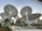 Innovative, large solar panel structures designed like futuristic trees, situated in a landscaped area with various plants and a pathway. The scene includes people walking, and the architecture emphasizes a modern, sustainable design.