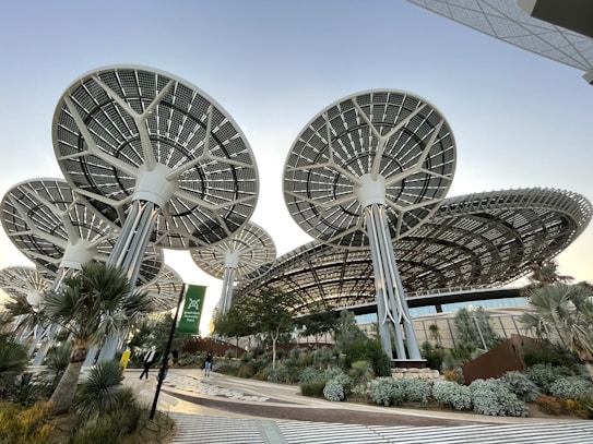 Innovative, large solar panel structures designed like futuristic trees, situated in a landscaped area with various plants and a pathway. The scene includes people walking, and the architecture emphasizes a modern, sustainable design.