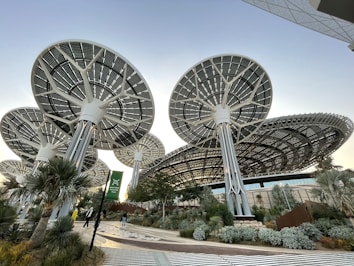 Innovative, large solar panel structures designed like futuristic trees, situated in a landscaped area with various plants and a pathway. The scene includes people walking, and the architecture emphasizes a modern, sustainable design.