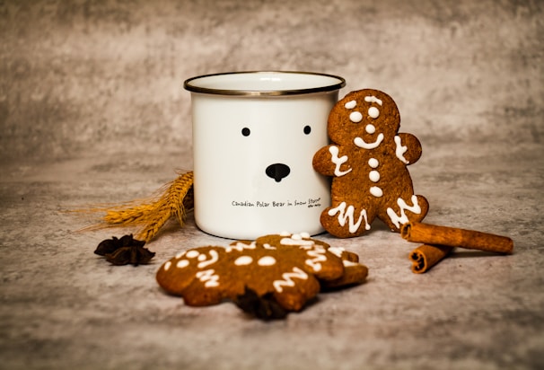 On a textured surface, there is a white mug featuring a simplistic bear face design. Next to the mug are gingerbread cookies, one upright and the other lying flat. Both cookies are decorated with white icing. Surrounding the items are pieces of cinnamon sticks and wheat spikes.