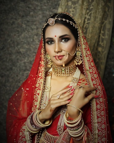 A bride adorned in traditional Indian wedding attire. She is wearing a red lehenga with intricate gold embroidery and matching jewelry, including a heavy gold necklace, bangles, and an ornate headpiece. Her hands show detailed henna designs, and she has a delicate nose ring. The makeup is elegant, emphasizing her eyes and lips.