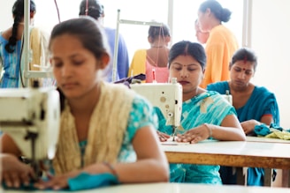 A group of women are working in a sewing room. The women in the foreground are seated at tables using sewing machines, while others are standing and working in the background. They are dressed in colorful clothing, mainly blue and orange. The overall scene conveys a busy and industrious environment.