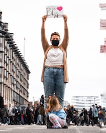 A person wearing a mask holds a sign that reads 'Black Lives Matter' with a heart symbol, standing on a street with buildings in the background. A child is crouched at their feet, looking to the side. People and bicycles are visible in the background, suggesting a protest or gathering.