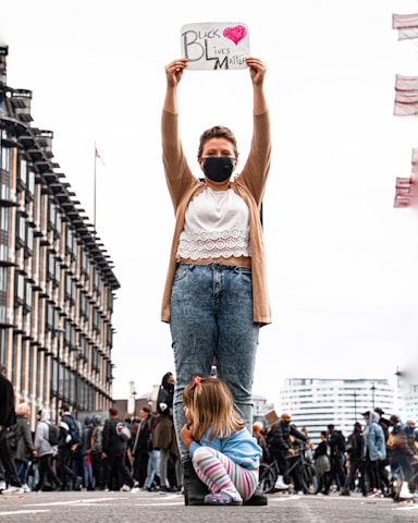 A person wearing a mask holds a sign that reads 'Black Lives Matter' with a heart symbol, standing on a street with buildings in the background. A child is crouched at their feet, looking to the side. People and bicycles are visible in the background, suggesting a protest or gathering.