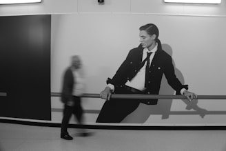 A blurred motion captures an older man walking by a large, black and white fashion poster. The poster features a young man in a suit with a stylish demeanor, leaning on a railing. The scene is set in what appears to be an indoor space with smooth flooring and a stark, modern aesthetic.
