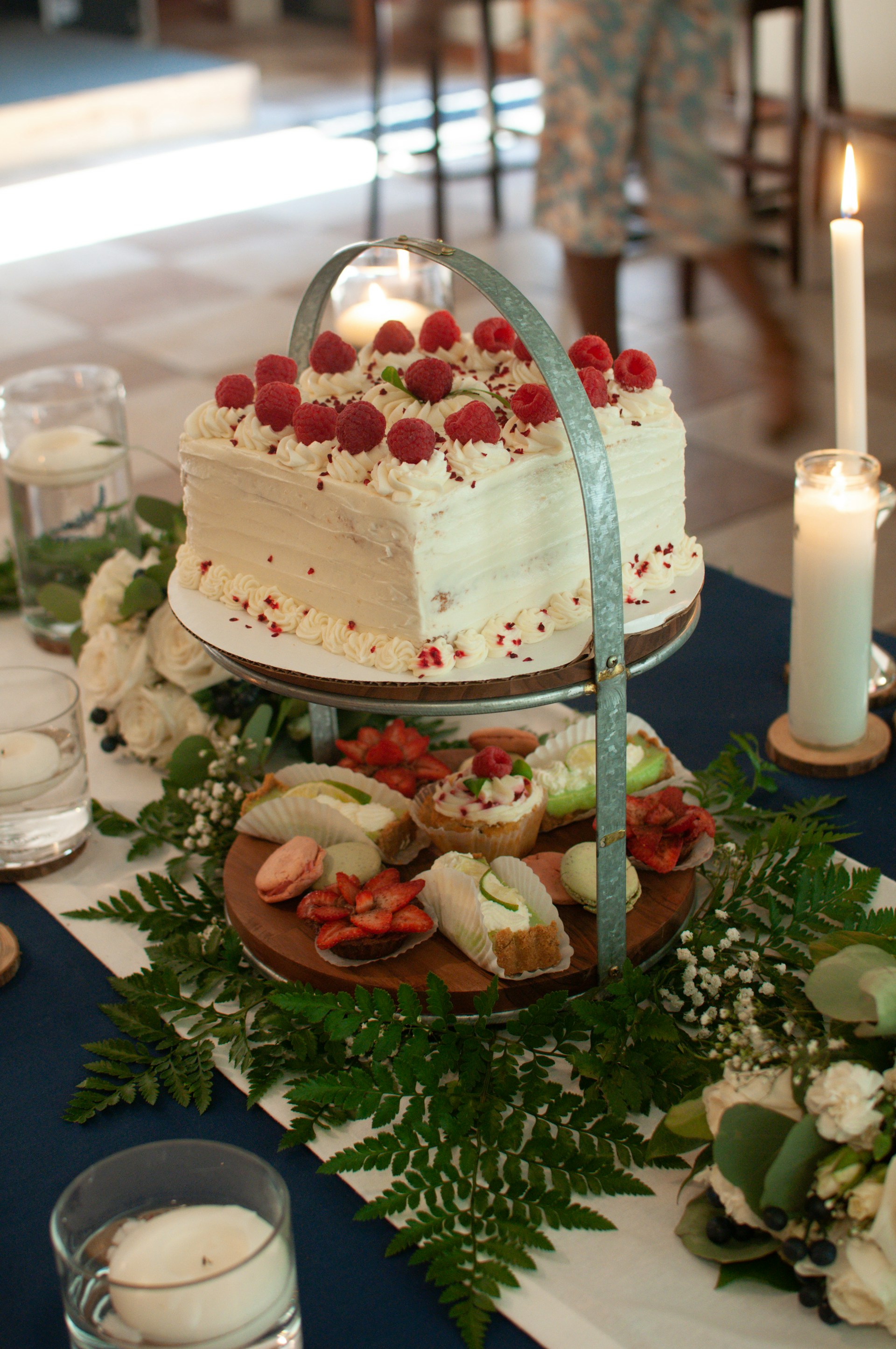 A white frosted cake adorned with fresh raspberries placed on a two-tiered stand. Below the cake, an assortment of desserts including macarons, fruit slices, and pastries are displayed on the lower tier. The setup is decorated with green ferns and white flowers, adding a natural touch. Candles in glass holders are placed around the table setting, enhancing the ambiance.