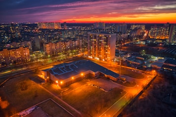 Aerial view of a cityscape at dusk with numerous high-rise apartment buildings. The sky is a vibrant orange and purple, contrasting with the darkening city below. Streetlights and windows illuminate the scene, creating a warm glow. In the foreground, a large building surrounded by green space is lit up brightly.