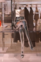 A metallic mannequin torso and legs are displayed in a store window, with a white handbag adorned with a gold chain placed on top of the hips. The background includes blurred clothing racks and various garments hanging, adding depth to the scene.