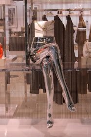 A metallic mannequin torso and legs are displayed in a store window, with a white handbag adorned with a gold chain placed on top of the hips. The background includes blurred clothing racks and various garments hanging, adding depth to the scene.