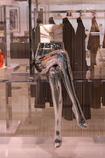A metallic mannequin torso and legs are displayed in a store window, with a white handbag adorned with a gold chain placed on top of the hips. The background includes blurred clothing racks and various garments hanging, adding depth to the scene.