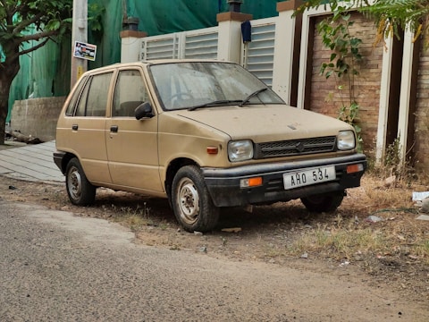 A dusty, old compact car parked beside a paved road. The car has a beige color and is showing signs of wear and age. It is situated in an urban environment with a misaligned number plate that reads 'AHO 534'. There is vegetation and a brick wall in the background, along with a tree on one side.
