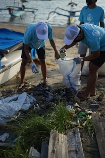 Several individuals wearing blue Ocean-themed shirts and white caps are actively collecting trash on a sandy beach. They are sorting litter into a large white bag, surrounded by various discarded items like plastic bottles and debris. In the background, small boats are moored on the water, suggesting a coastal cleanup effort.