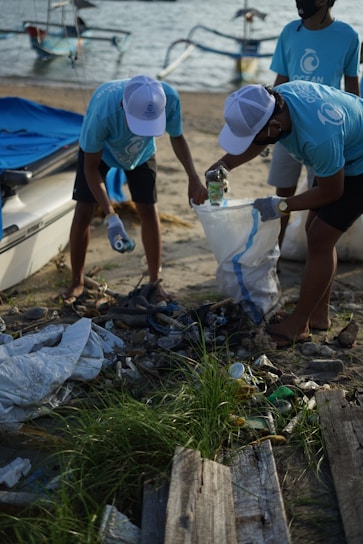 Several individuals wearing blue Ocean-themed shirts and white caps are actively collecting trash on a sandy beach. They are sorting litter into a large white bag, surrounded by various discarded items like plastic bottles and debris. In the background, small boats are moored on the water, suggesting a coastal cleanup effort.