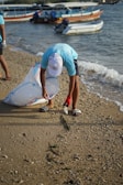 A person wearing a light blue shirt and white cap is picking up trash on a sandy beach. They are holding a large, transparent bag and are bending down to collect a plastic bottle. In the background, several boats are docked in the water.