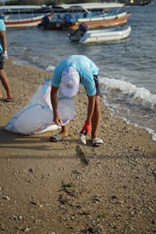 A person wearing a light blue shirt and white cap is picking up trash on a sandy beach. They are holding a large, transparent bag and are bending down to collect a plastic bottle. In the background, several boats are docked in the water.
