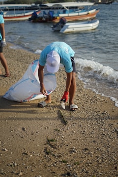 A person wearing a light blue shirt and white cap is picking up trash on a sandy beach. They are holding a large, transparent bag and are bending down to collect a plastic bottle. In the background, several boats are docked in the water.