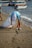 A person wearing a light blue shirt and white cap is picking up trash on a sandy beach. They are holding a large, transparent bag and are bending down to collect a plastic bottle. In the background, several boats are docked in the water.
