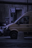A delivery van is parked on the street next to a smaller car. The van has the words 'Service Partner of DHL' on its side. There is a parking meter and a sign labeled 'PARCHIMETRO' with an arrow pointing to 'TICKET'. The scene takes place in a dimly lit urban environment.