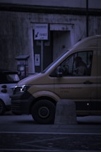 A delivery van is parked on the street next to a smaller car. The van has the words 'Service Partner of DHL' on its side. There is a parking meter and a sign labeled 'PARCHIMETRO' with an arrow pointing to 'TICKET'. The scene takes place in a dimly lit urban environment.