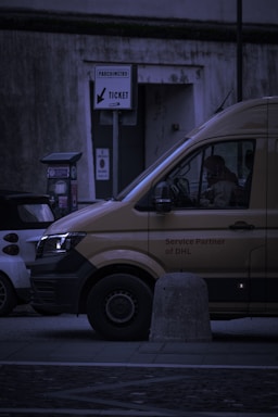 A delivery van is parked on the street next to a smaller car. The van has the words 'Service Partner of DHL' on its side. There is a parking meter and a sign labeled 'PARCHIMETRO' with an arrow pointing to 'TICKET'. The scene takes place in a dimly lit urban environment.