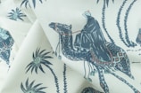 A fabric with a printed pattern featuring a person dressed in traditional clothing riding a camel. The design includes palm trees and decorative motifs in an artistic style, using a limited color palette.