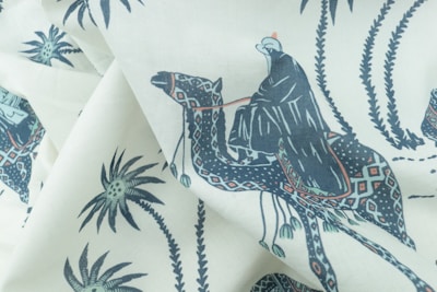 A fabric with a printed pattern featuring a person dressed in traditional clothing riding a camel. The design includes palm trees and decorative motifs in an artistic style, using a limited color palette.